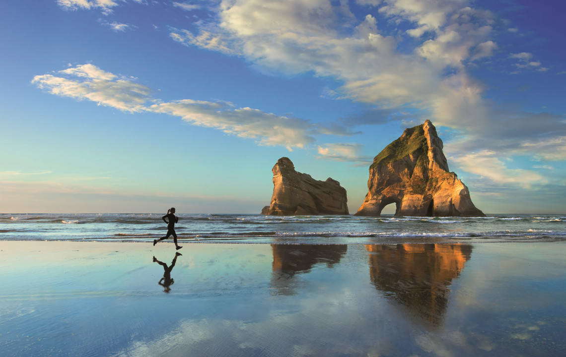 Eine Frau joggt am Strand. Der Morgenhimmel und eine Felsformation im Hintergrund spiegeln sich im Wasser