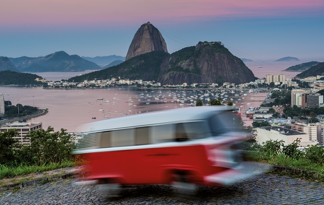 Verschwommene Aufnahme eines roten VW Bulli in voller Fahrt. Im Hintergrund ist der Zuckerhut und der Hafen von Rio de Janeiro zu sehen.