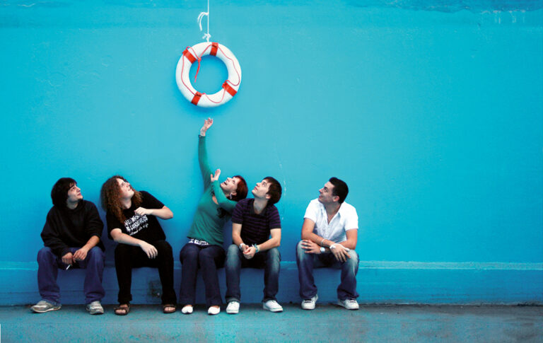 Eine Gruppe junger Menschen sitzt vor einer blauen Wand. Über ihnen hängt ein Rettungsring, zu dem sie aufschauen