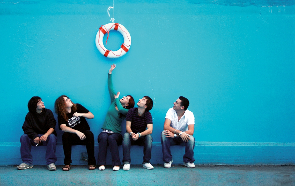Eine Gruppe junger Menschen sitzt vor einer blauen Wand. Über ihnen hängt ein Rettungsring, zu dem sie aufschauen