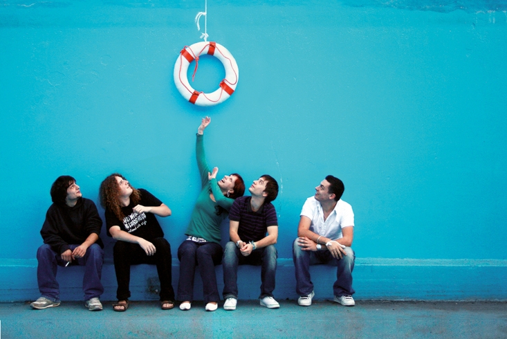 Eine Gruppe junger Menschen sitzt vor einer blauen Wand. Über ihnen hängt ein Rettungsring, zu dem sie aufschauen. Link führt zur Leseprobe Die innere WG
