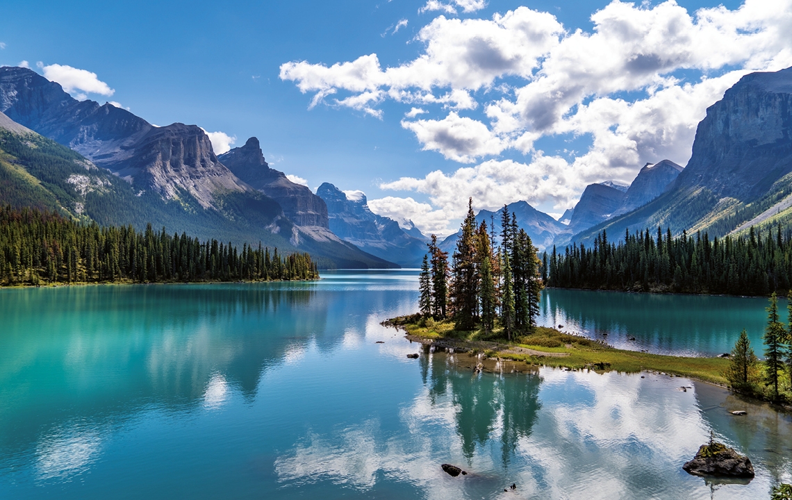 Panoramaaufnahme eines Gebirgssees. Im strahlend blauen Wasser spiegelt sich die Umgebung und der Himmel