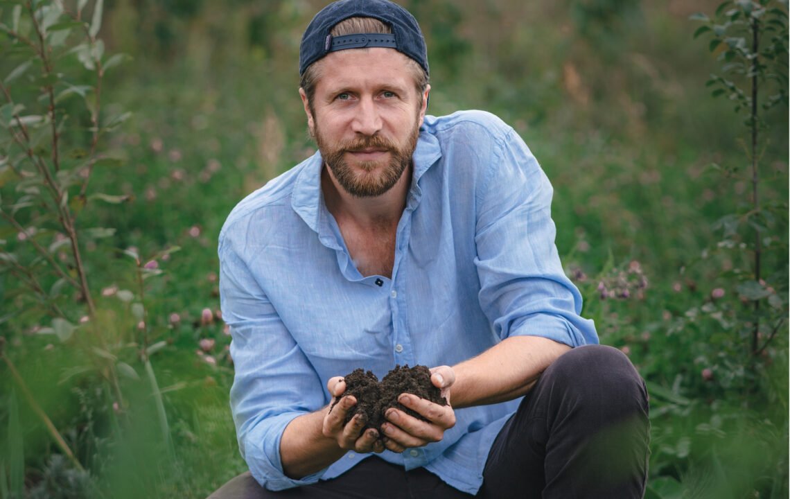 Landwirt Benedikt Bösel kniet in einem Feld und hält fruchtbare Erde in seinen Händen